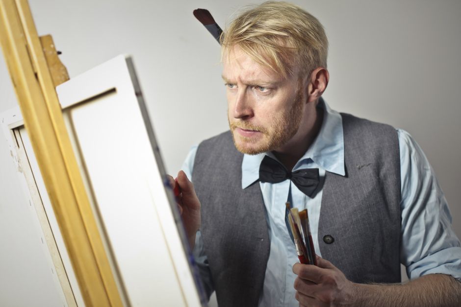 Focused male artist in elegant outfit creating artwork using canvas and easel while standing in workshop with paintbrushes